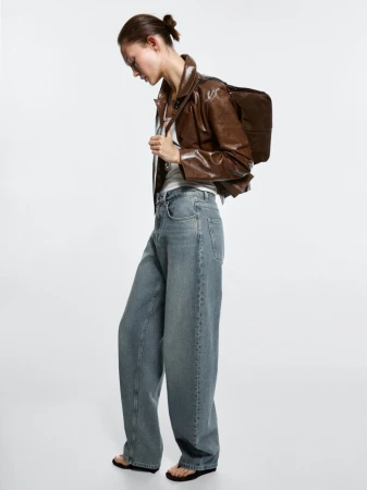 A young woman in a brown jacket and baggy jeans stands against a white background in a fashion shot. She's carrying a brown bag.