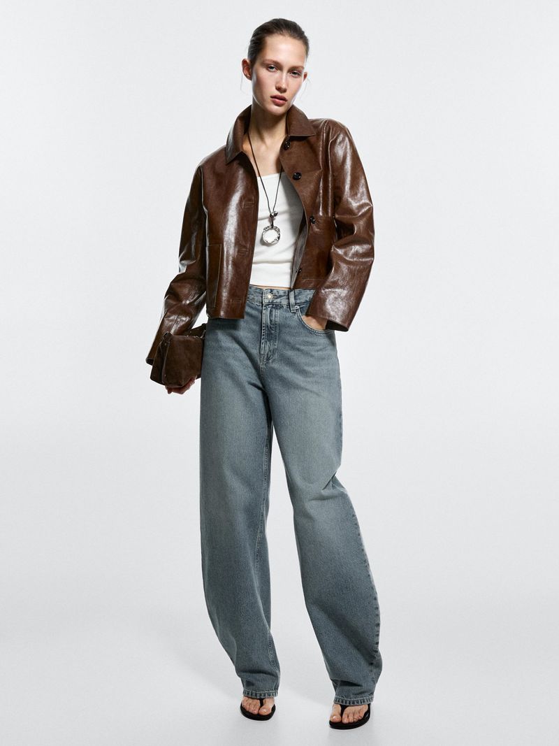 A woman stands in a photo studio wearing a brown jacket, light blue jeans, a white tank top, and black flip-flops.