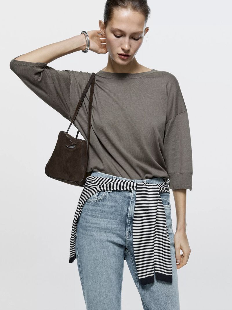 Fashionable adult woman poses in light blue jeans and a gray top with a brown handbag over her shoulder in a studio shot.