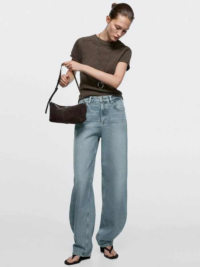 Model wearing brown knit top and light wash denim jeans holding a dark brown handbag against a white background.