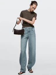 Model wearing brown knit top and light wash denim jeans holding a dark brown handbag against a white background.