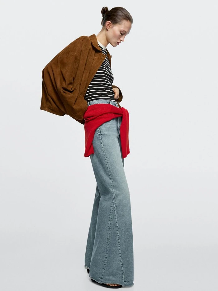 Model wearing a brown jacket, striped top, and wide-leg denim jeans against a plain white seamless studio background.