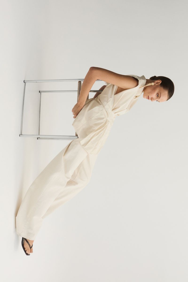 Model wearing a cream sleeveless top and matching trousers leaning against a metal chair on white background