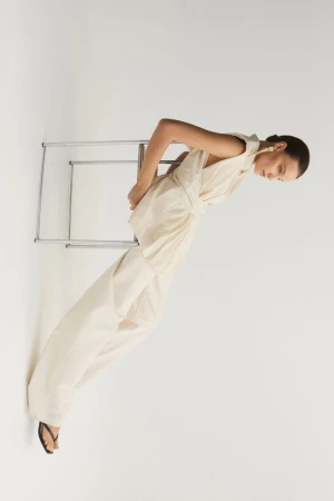 Model wearing a cream sleeveless top and matching trousers leaning against a metal chair on white background