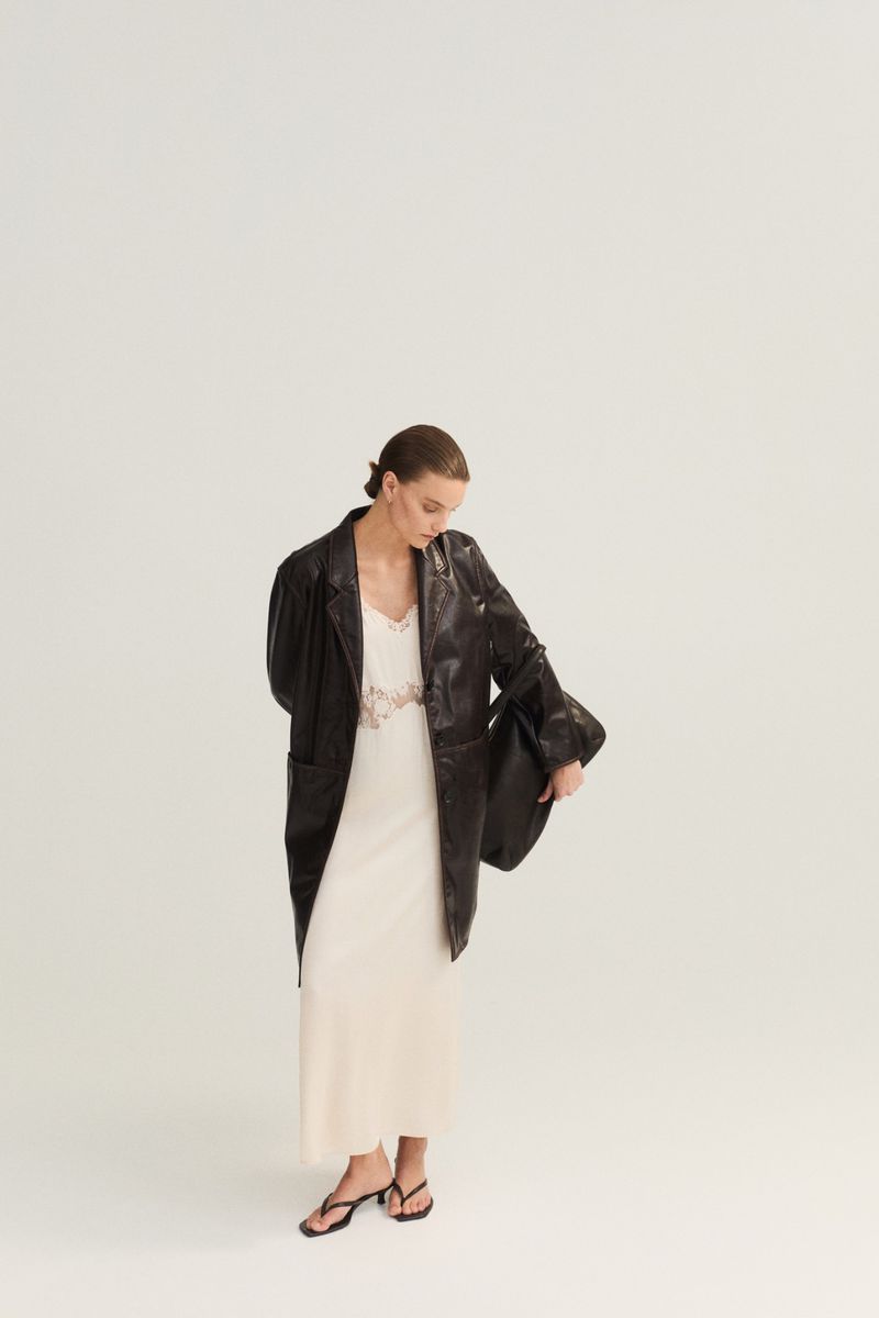 A stylish adult woman stands indoors in a cream dress and black leather blazer, holding a black bag against a plain background.