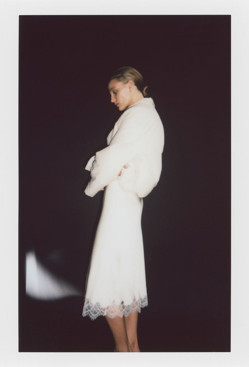 Model in a white dress with lace hem standing against a dark backdrop for the H&M SIMPLICITÉ project.