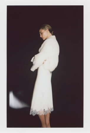 Model in a white dress with lace hem standing against a dark backdrop for the H&M SIMPLICITÉ project.