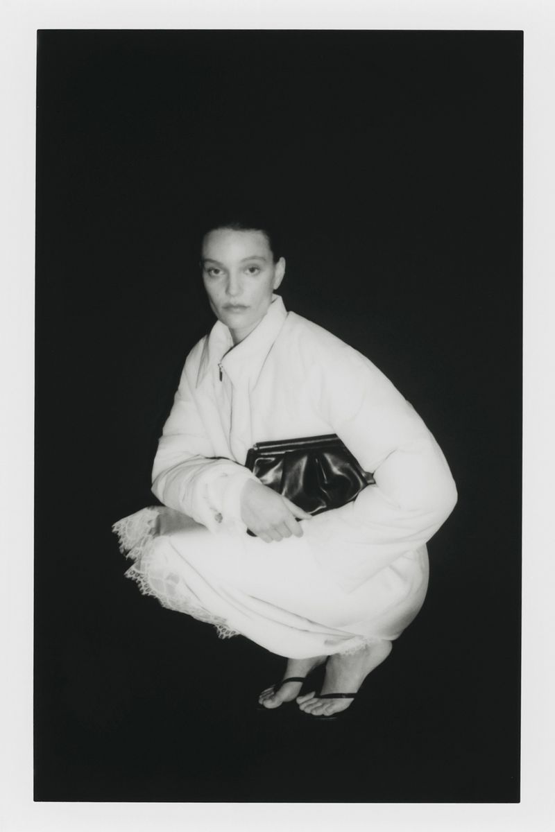 A woman squats in a photo studio, modelling fashion with a black clutch bag against a dark backdrop, evoking monochrome elegance.
