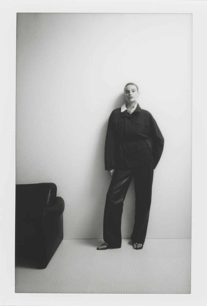 Model standing against a plain white wall wearing a dark jacket and matching trousers beside a black armchair.