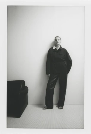 Model standing against a plain white wall wearing a dark jacket and matching trousers beside a black armchair.