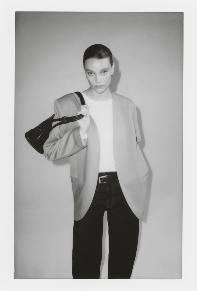 Model in blazer holding a black handbag against a grey studio backdrop for H&M SIMPLICITÉ