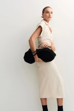 A model in a cream-colored midi dress holds a large black woven handbag against a plain, light-colored background.