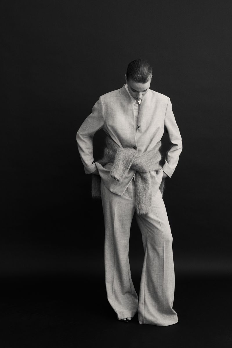 Woman poses in a grey oversized suit with a sweater tied around her waist in a studio shot against a dark backdrop.