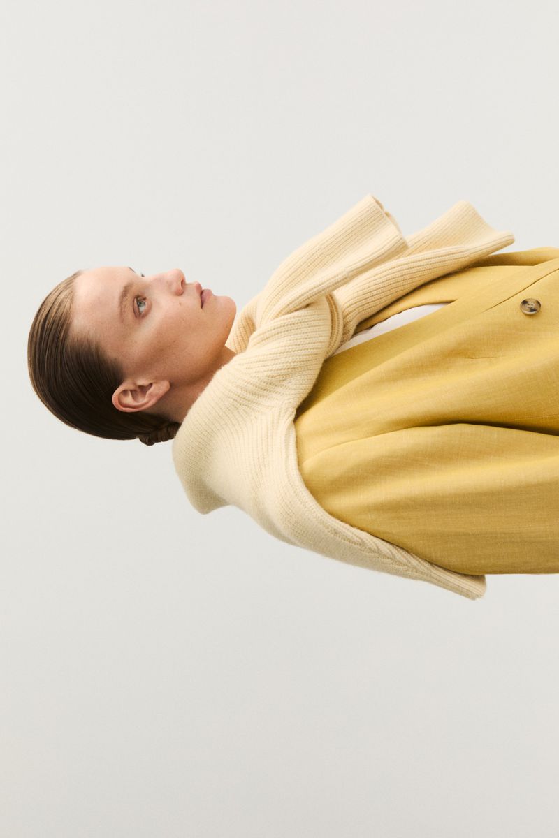 A woman in a yellow blazer and cream-colored sweater poses against a white background, showcasing a minimalist fashion aesthetic.
