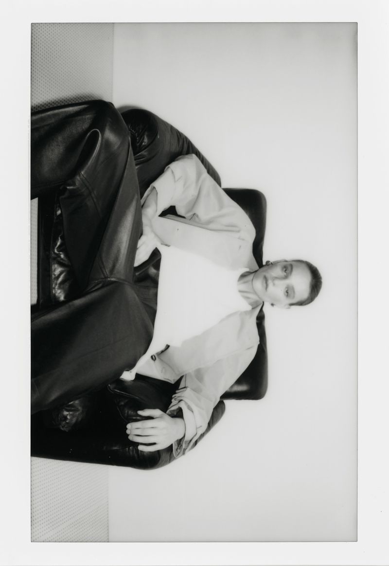Monochromatic portrait of a female fashion model reclining in a black leather chair in a bright indoor setting, styled in a modern outfit.