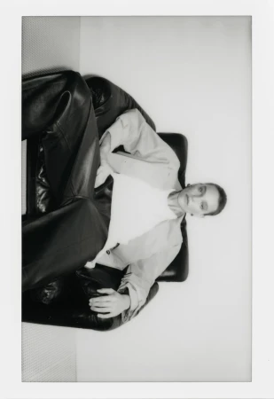 Monochromatic portrait of a female fashion model reclining in a black leather chair in a bright indoor setting, styled in a modern outfit.