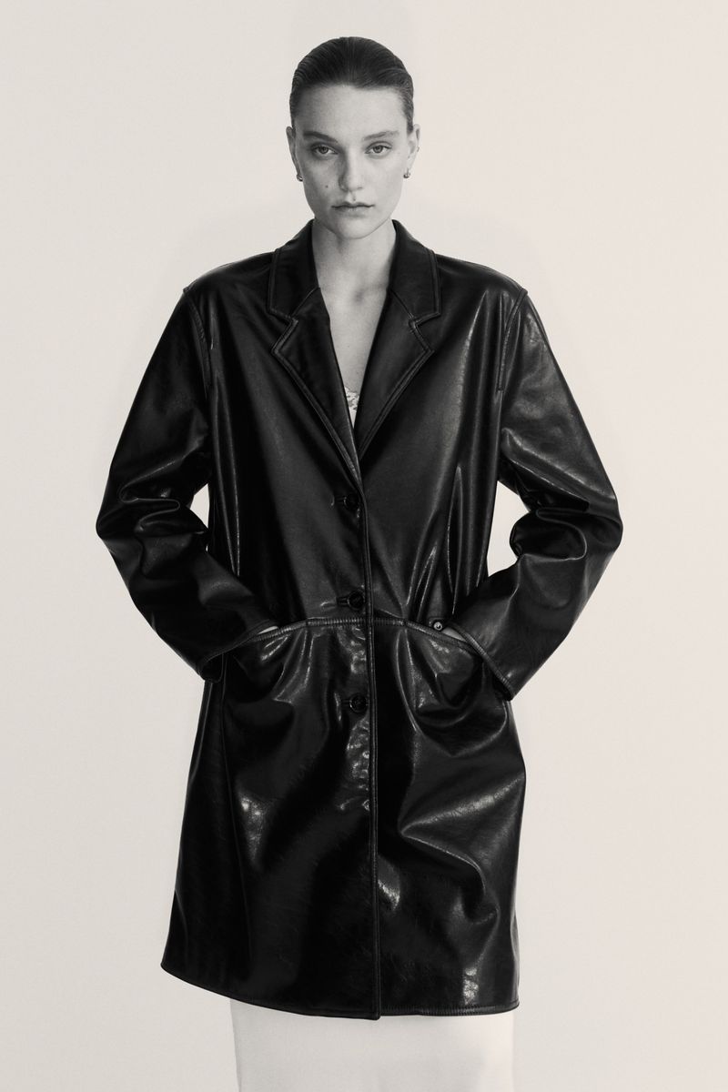 A person with hands in pockets wears a shiny black knee-length coat in a studio setting with soft lighting, shown in a portrait orientation.