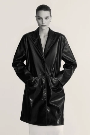A person with hands in pockets wears a shiny black knee-length coat in a studio setting with soft lighting, shown in a portrait orientation.