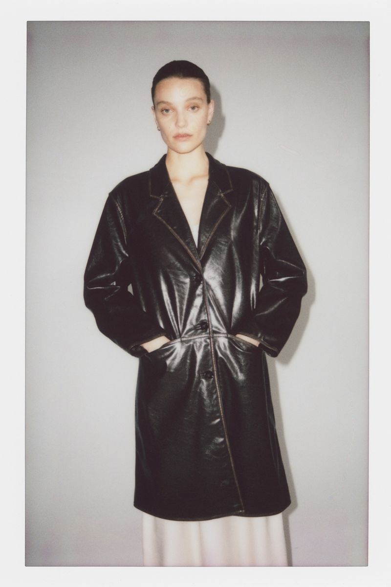 Woman in a black patent leather trench coat, hands in her pockets, standing indoors against a neutral studio backdrop.