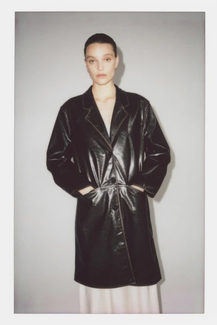 Woman in a black patent leather trench coat, hands in her pockets, standing indoors against a neutral studio backdrop.