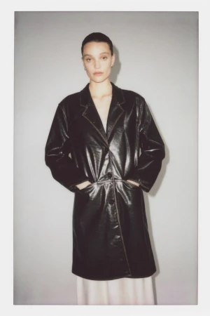 A woman wearing a black leather coat with hands in her pockets stands against a neutral grey wall.