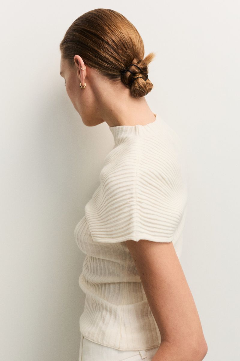 Back view of a woman with her hair in a bun wearing a ribbed, short-sleeve, mock-neck sweater against a neutral backdrop.