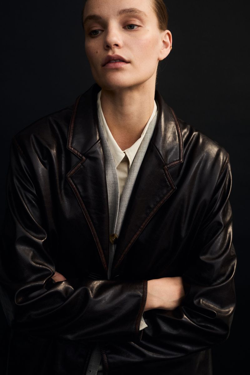 Portrait of a young woman wearing a brown leather jacket and light shirt, standing indoors against a dark background with her arms crossed.