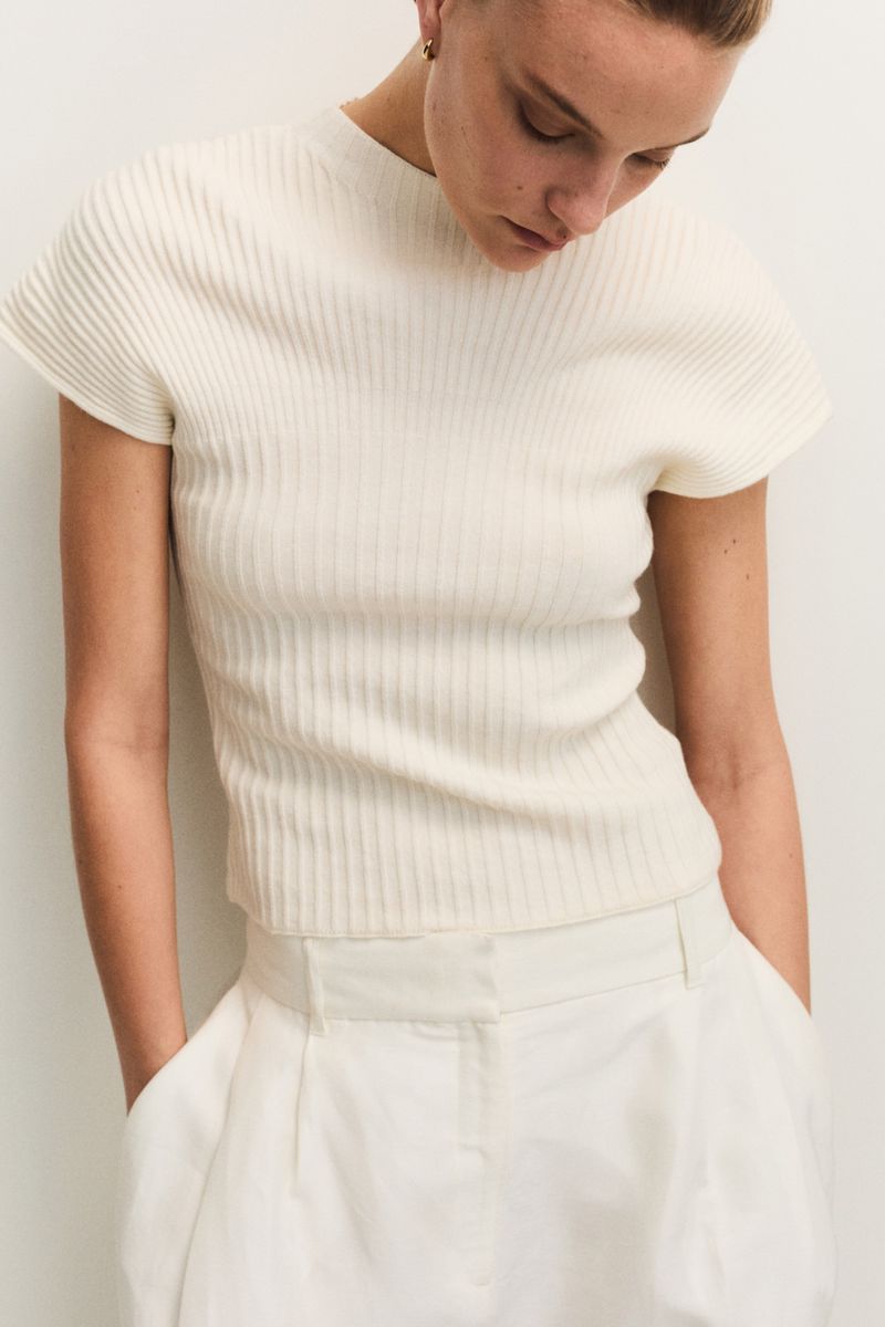 Close-up of a fair-skinned woman wearing a ribbed white knit top and white pants, hands in pockets, looking downward in a neutral indoor setting.