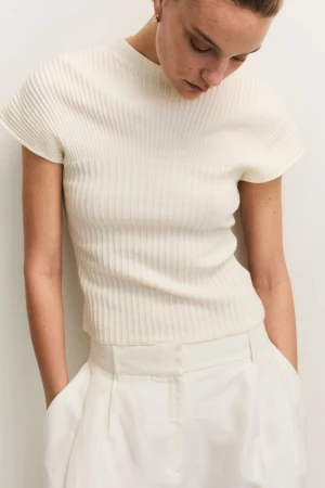 Close-up of a fair-skinned woman wearing a ribbed white knit top and white pants, hands in pockets, looking downward in a neutral indoor setting.