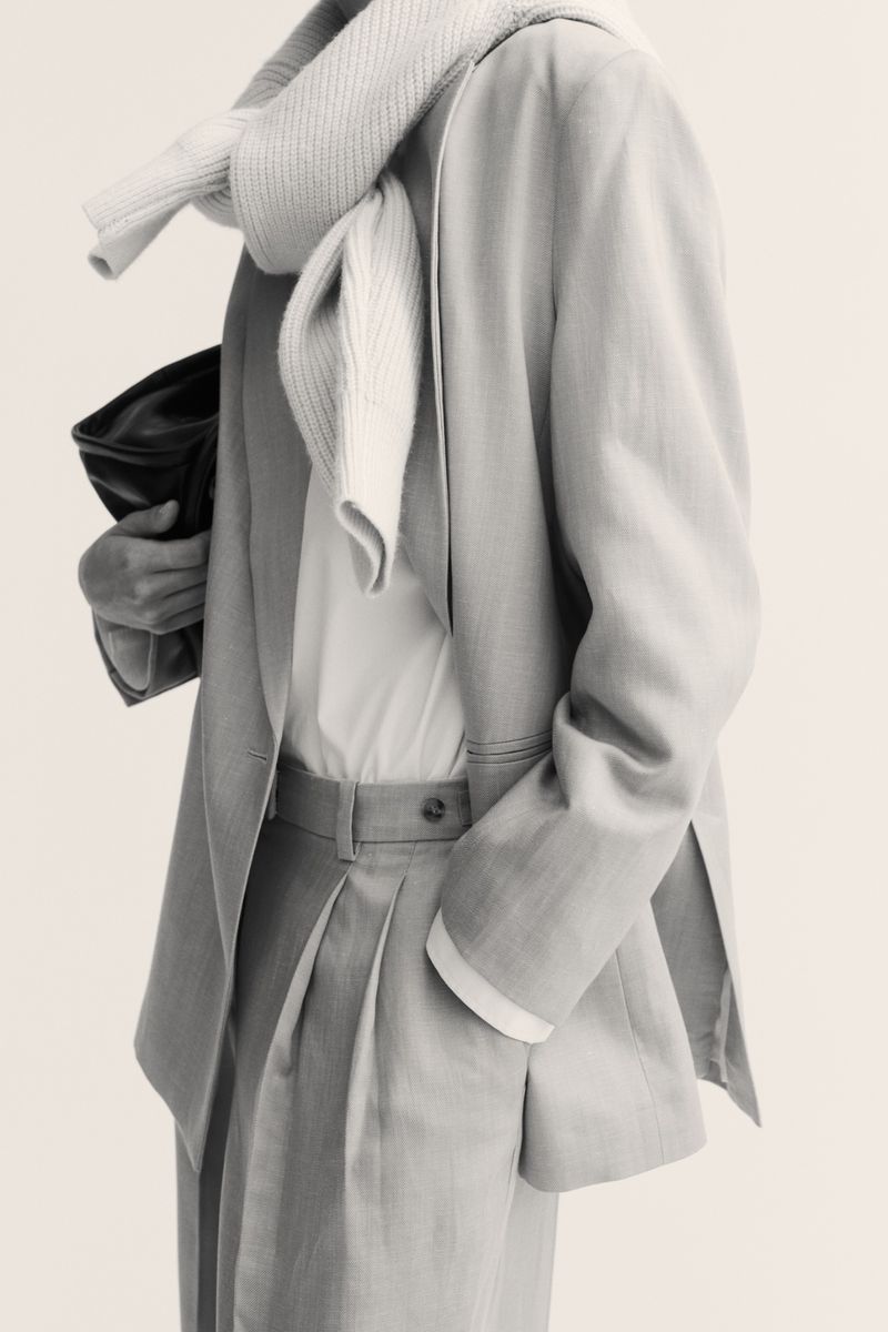 Fashion editorial featuring a woman wearing a tailored blazer, trousers, and cream scarf, posing in a minimalist indoor setting.