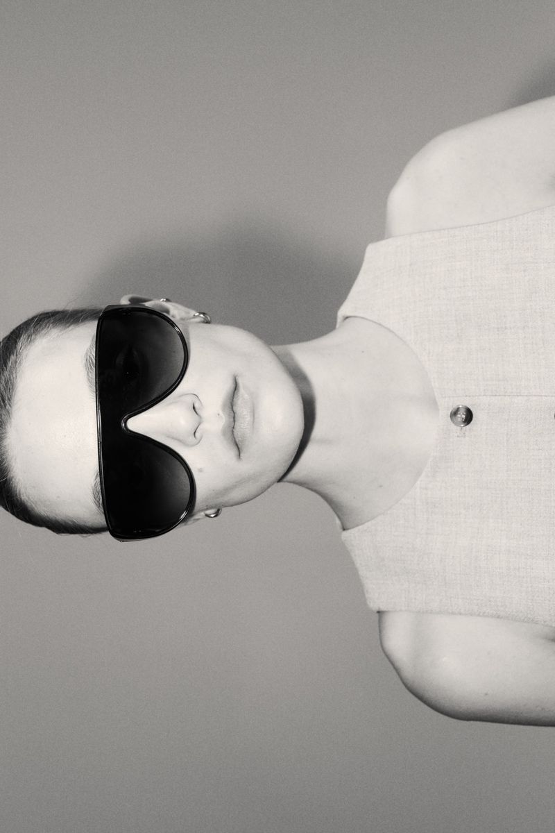 Model wearing dark oversized sunglasses and a sleeveless top against a grey seamless studio background.
