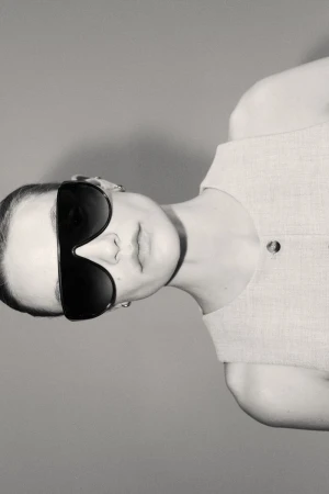 Model wearing dark oversized sunglasses and a sleeveless top against a grey seamless studio background.