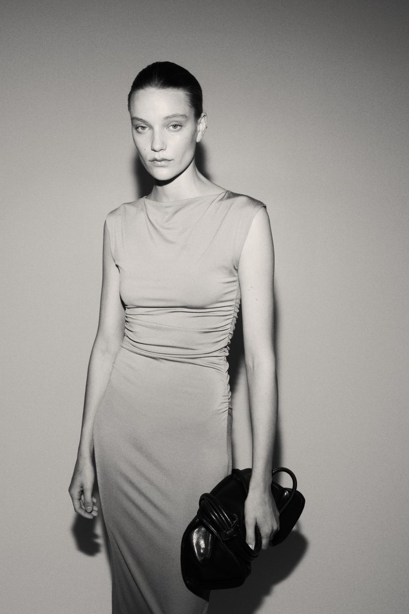 Monochrome portrait of a woman in a fitted dress holding a structured leather handbag against a plain background in a studio.