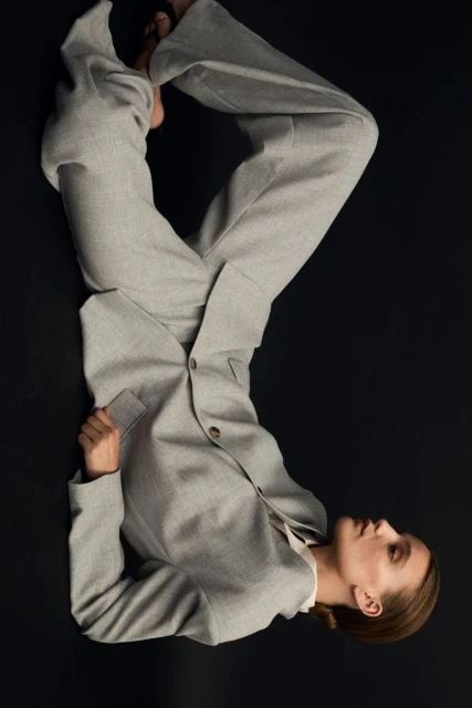 Fashion shot of an adult woman lying on a black surface wearing a grey suit jacket and wide-leg pants, set against a dark backdrop.