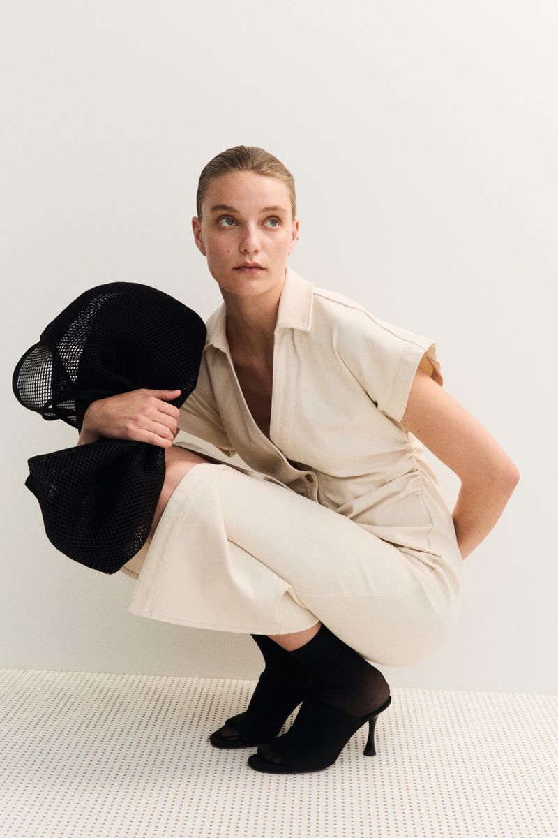 Woman poses in collared beige jumpsuit with black accessories on a white patterned floor, creating a minimalist fashion editorial.