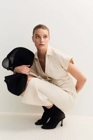 Woman poses in collared beige jumpsuit with black accessories on a white patterned floor, creating a minimalist fashion editorial.
