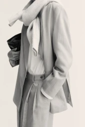 Model in grey tailored blazer wearing a white sweater draped over shoulders and carrying a black clutch bag