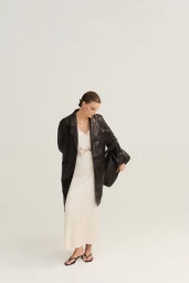 Model in a long dark leather coat over a cream slip dress holding a black handbag against a white studio background.