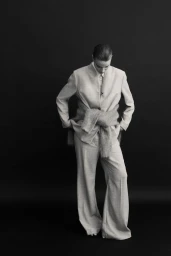 Model in a light-coloured suit and fluffy sweater tied around the waist standing against a plain black background.