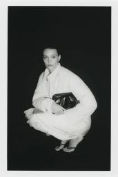 Model in white jacket and skirt holding a black clutch against a dark background