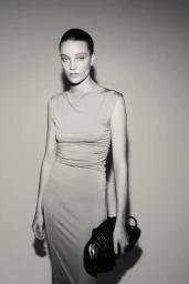 Model in sleeveless midi dress holding a black handbag against a neutral grey background