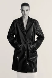 Model wearing a black leather coat with hands in pockets against a neutral backdrop