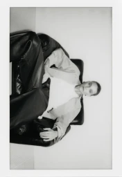Model seated in a black leather armchair wearing dark trousers and a light jacket against a white studio background.