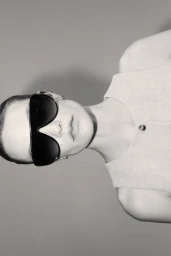 Model wearing dark oversized sunglasses and a sleeveless top against a grey seamless studio background.
