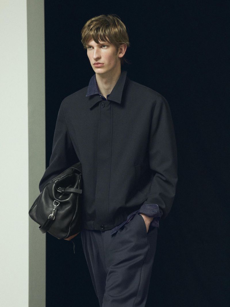 Male model wearing a dark jacket carrying a black leather holdall bag against a dark background for Massimo Dutti