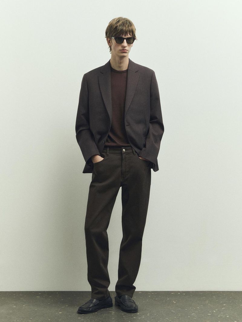 A stylish adult male in a brown suit and sunglasses stands against a light wall, showcasing fashion and simple studio photography.
