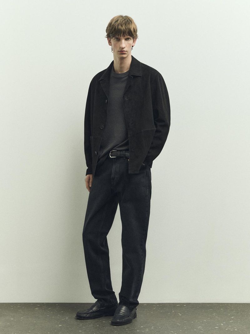A stylish adult male stands in a studio wearing a black jacket, gray t-shirt, and dark jeans against a neutral background for a fashion shoot.