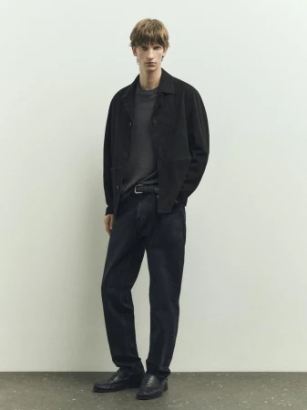 A stylish adult male stands in a studio wearing a black jacket, gray t-shirt, and dark jeans against a neutral background for a fashion shoot.