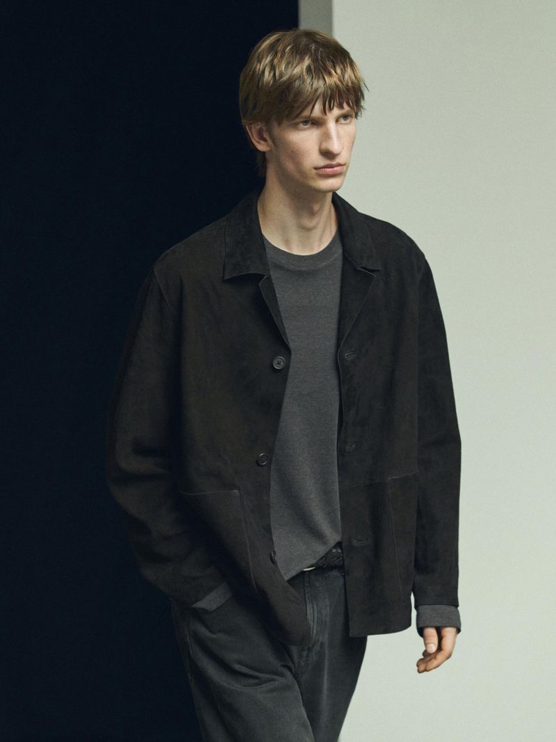 Fashion portrait of a blonde male model in a black jacket and grey t-shirt posing in a photo studio with a two-tone background.
