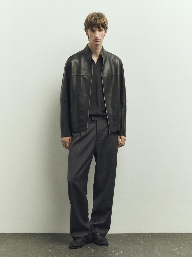 Man in black leather jacket, dark shirt, and grey slacks standing in a neutral setting in a fashion editorial shot. 